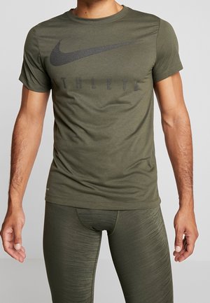 Man draagt een olijfgroen Nike T-shirt met de tekst "ATHLETE" en bijpassende getextureerde sportleggings, staand tegen een effen achtergrond.