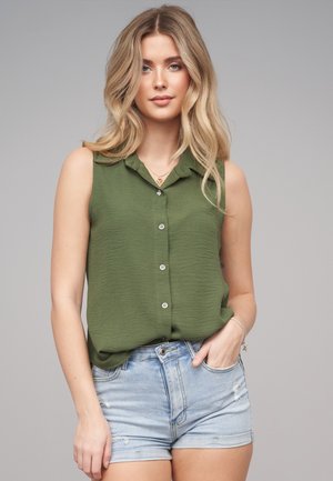 Blonde Frau trägt ärmellose olivgrüne Bluse mit Knöpfen und hellblaue, ausgewaschene Jeansshorts, steht vor schlichtem grauem Hintergrund.
