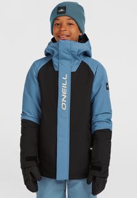 Blauwe en zwarte geïsoleerde jas met een hoge kraag, voorzien van een ritssluiting aan de voorkant en logo-details, aangevuld met zwarte handschoenen en een blauwe beanie.