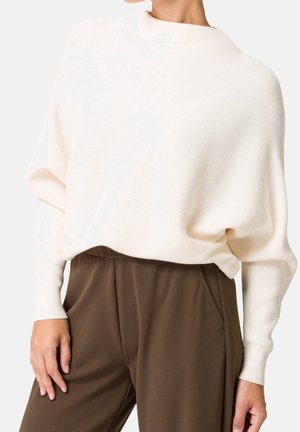 Femme portant un pull ample côtelé de couleur blanc cassé à manches longues et un pantalon marron taille haute.