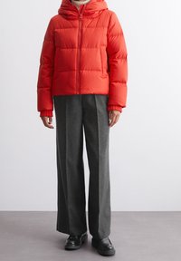 Czerwona kurtka puchowa z kapturem, z zamkiem błyskawicznym i ściśniętymi mankietami, w połączeniu z szarymi szerokimi spodniami i czarnymi butami.