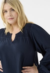 Tmavě modrá blůza s texturovanou tkaninou, která má nařasený výstřih, dlouhé rukávy se sbíranými manžetami a volný střih.