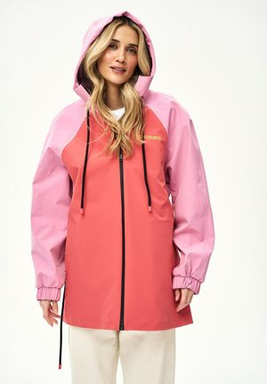 Kobieta o długich blond włosach, ubrana w dwukolorową różowo-koralową kurtkę przeciwdeszczową z kapturem, z czarnymi sznurkami i zamkiem błyskawicznym, stojąca na białym tle.