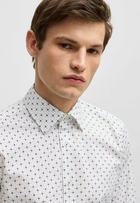Witte button-up shirt met een geometrisch blauw patroon, gestructureerde kraag en textiele stof, met zichtbare knopen aan de voorkant.
