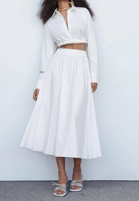 Chemisier blanc court avec des manches longues et un col haut associé à une jupe midi blanche fluide, présentant des détails froncés et une texture lisse.
