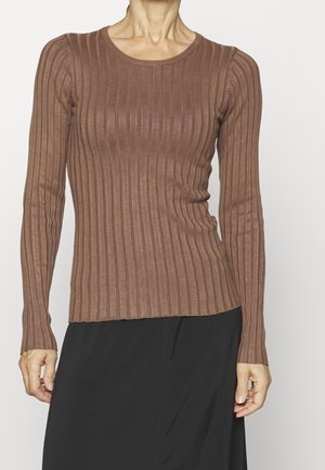 Vrouw draagt een nauwsluitende, langärmelig, geribde bruine trui gecombineerd met een zwarte rok tegen een effen witte achtergrond.