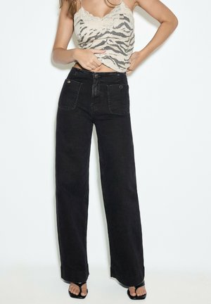 Kvinde iført sorte jeans med vide ben og forlommer, en tigerstribet spidskantet camisole og sorte sandaletter med hæl.