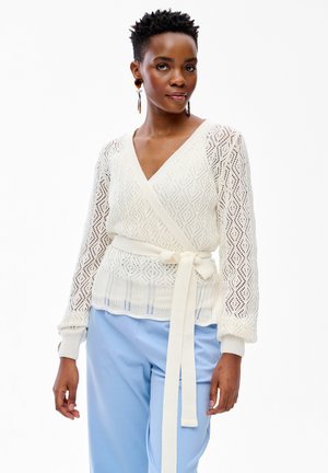 Woman wearing white knit wrap sweater with belt and light blue pants, standing with neutral expression on a white background.