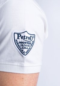 Bílá košile s krátkým rukávem, která má na horním rukávu modrou vyšitou nášivku ve tvaru štítu se nápisem "Petrol Industries" a detaily o místě.