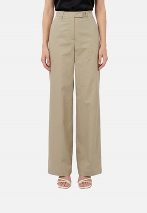 Donna che indossa pantaloni beige a gamba larga e vita alta abbinati a sandali bianchi con cinturini e una maglia nera, in piedi davanti a uno sfondo semplice.