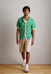 Grøn kortærmet skjorte med hvide geometriske mønstre, parret med beige shorts. Forstærkede syninger og lette sneakers fuldender looket.