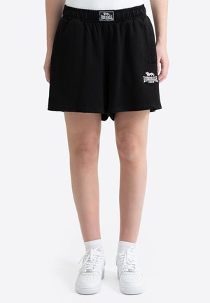 Persona que lleva puestos pantalones cortos negros Lonsdale London, calcetines blancos y zapatillas blancas, de pie con las piernas visibles desde la mitad del muslo hacia abajo.