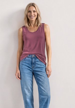 Lächelnde Frau mit schulterlangem blondem Haar, die ein ärmelloses mauvefarbenes Oberteil und hellblaue Jeans trägt, steht vor einer schlichten hellen Wand.