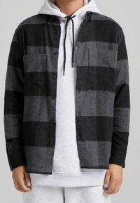 Man bär en svart och grå rutad skjorta med knappar över en ljusgrå hoodie med svarta snören, tillsammans med ljusgrå byxor.