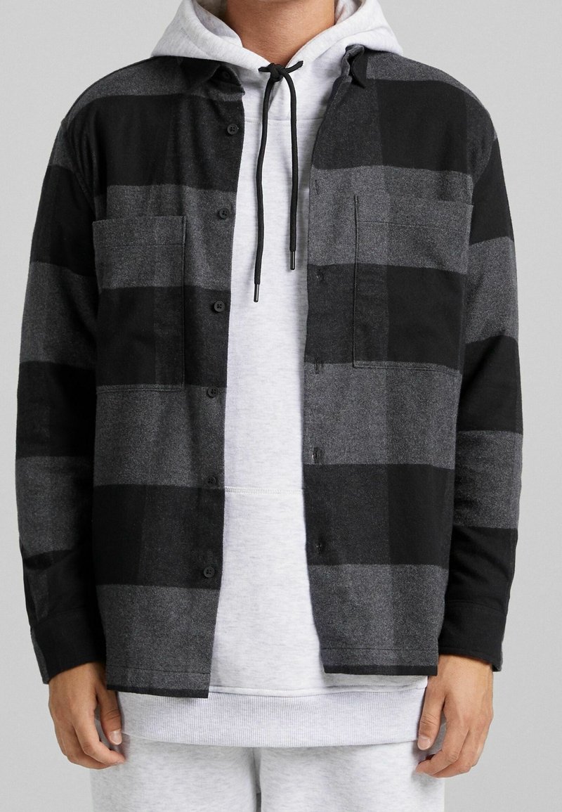 Man bär en svart och grå rutad skjorta med knappar över en ljusgrå hoodie med svarta snören, tillsammans med ljusgrå byxor.