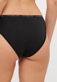Culotte en coton noir avec des bordures en dentelle, dotée d'une taille élastique marquée "TOMMY HILFIGER." Texture lisse et design classique offrant une couverture complète.