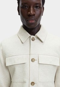 Jasnobeżowa wełniana kurtka z kołnierzem, dwoma przednimi kieszeniami i brązowymi guzikami, prezentująca miękką teksturę i dopasowany krój.