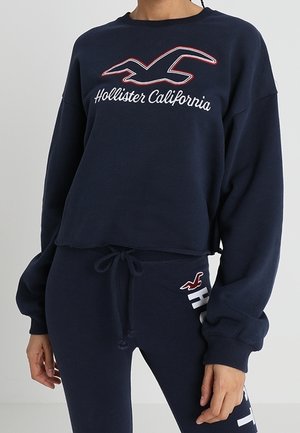 Sudadera azul marino de manga larga y corta con texto "Hollister California" y logotipo de gaviota, combinada con pantalones deportivos a juego con cordón.