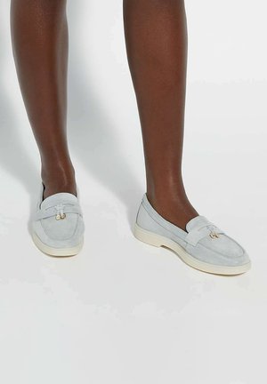 Light blue suede loafers with cream rubber soles, featuring a decorative bow and gold-tone hardware detail at the front.
