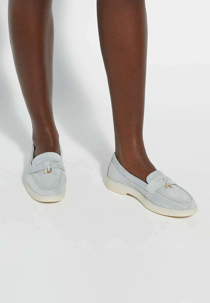 Light blue suede loafers with cream rubber soles, featuring a decorative bow and gold-tone hardware detail at the front.