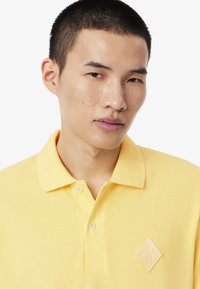 Polo gialla con piccolo logo sul petto, caratterizzata da un tessuto strutturato e un colletto tradizionale. Due bottoni all'altezza del collo.