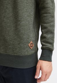 Donkergroene sweatshirt met een gestructureerde afwerking, uitgerust met een bruine, ruitvormige logo patch aan de onderzijde links, met ribgebreide manchetten en zoom.