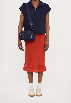 Maglietta a maniche corte blu navy, gonna di raso rossa con orlo a volant, borsa a tracolla blu navy con cerniere e sneakers bianche con suole chiare.