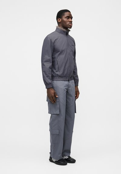 Γκρι ελαφρύ μπουφάν με όρθιο γιακά, ελαστική μέση; συνδυάζεται με γκρι παντελόνι cargo με πολλαπλές τσέπες και μαύρα παπούτσια.