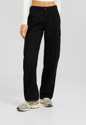 Kobieta w czarnych spodniach cargo z klapkowymi kieszeniami i beżowych tenisówkach, stojąca na jednolitym szarym tle.