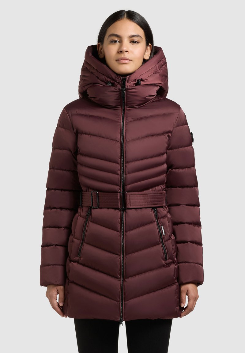 Bordo rdeča puhovka z zategnjenim pasom, visokim ovratnikom in veliko kapuco. Ponaša se z diagonalnim prešitjem in žepi z zadrgo spredaj za funkcionalnost.