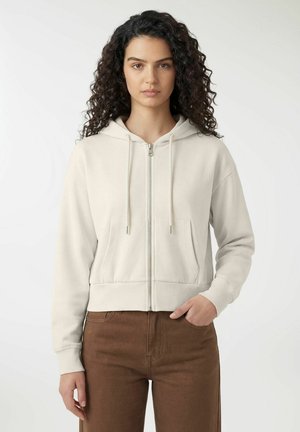 Młoda kobieta o kręconych włosach, ubrana w beżową bluzę rozpinaną na zamek i brązowe spodnie, stoi na tle jednolitego jasnego tła.