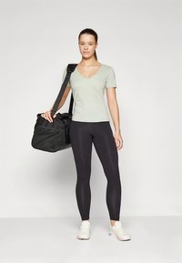 Lysegrøn T-shirt med V-hals, sorte leggings, hvide og lilla sneakers, holder en stor sort duffelbag med et mesh-lomme detalje.