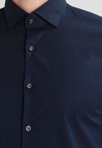 Mørkeblå skjorte med knapper, laget av et glatt materiale, klassisk krage og seks sølvknapper langs knappeåpningen. Enkel og elegant design.