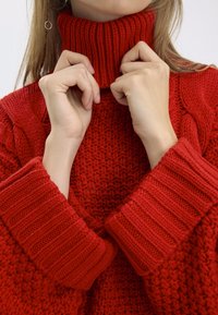 Pull rouge en tricot avec un col roulé côtelé montant. Les manches ont une texture côtelée épaisse, et le corps présente un motif tissé.