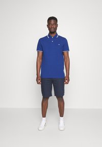 Mørkeblå polo skjorte med hvide detaljer, korte ærmer og knappet krave, parret med mørke shorts og hvide sneakers.