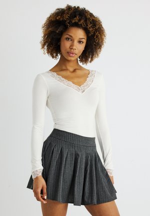 Γυναίκα με σγουρά μαλλιά, που φοράει λευκό μακρυμάνικο τοπ με δαντελένιο κέντημα και σκουρόχρωμη γκρι πλισέ φούστα με ρίγες.