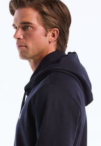 Haut à capuche bleu marine en tissu doux, avec une capuche à cordon de serrage, des coutures visibles le long des coutures, et un petit logo sur la poitrine.