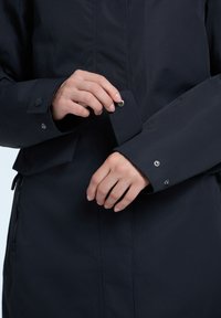 Giacca blu navy scuro con un bottone a scatto visibile sul polsino e chiusura del polsino regolabile. Presenta un tessuto liscio e dettagli di cucitura sottili.