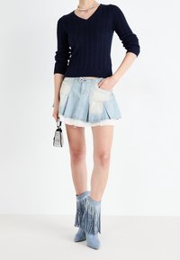 Kvinna i marinblå kabelstickad tröja, ljusblå plisserad denimminikjol, fransade blå ankelboots, håller en liten silverväska.