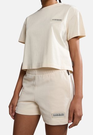 Person iført lyse beige crop top med korte ærmer og matchende shorts med Napapijri geografiske logo-patches på bryst og ben.