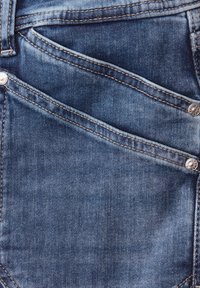 Gros plan sur un jean en denim bleu montrant deux poches avant superposées avec des rivets métalliques et des détails de couture visibles.