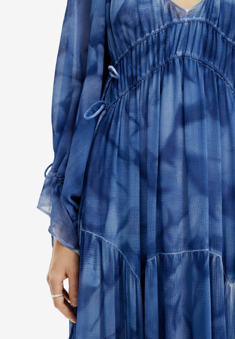 Blaues durchsichtiges Kleid mit einem gerafften Bund mit Bändern, langen Ärmeln und einem gestuften Rock. Der Stoff hat eine strukturierte, durchsichtige Qualität.