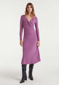 Vestido morado de manga larga con detalle de nudo en la parte delantera y una silueta fluida, combinado con botas negras hasta la rodilla. Textura suave, diseño de media longitud.