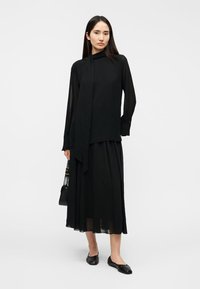 Μαύρο μπλούζα με μακριά μανίκια και κορδέλα στο λαιμό, φορεμένη πάνω από μια αέρινη μαύρη φούστα. Λείο ύφασμα με ελαφριά γυαλάδα, συνδυασμένο με μαύρα επίπεδα παπούτσια.