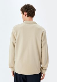 Beige pitkähihainen polo paita, jossa on kaulus, sileä pinta ja suora helma. Katsottuna takaa, ilman kuvioita tai koristeita.