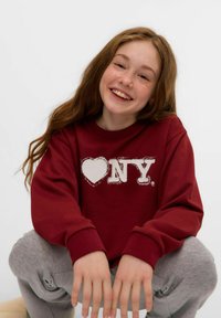 Vínová mikina s bílým potiskem "heart NYC", měkká textura, kulatý výstřih, nošena se šedými tepláky, modelka se usmívá a dřepí.