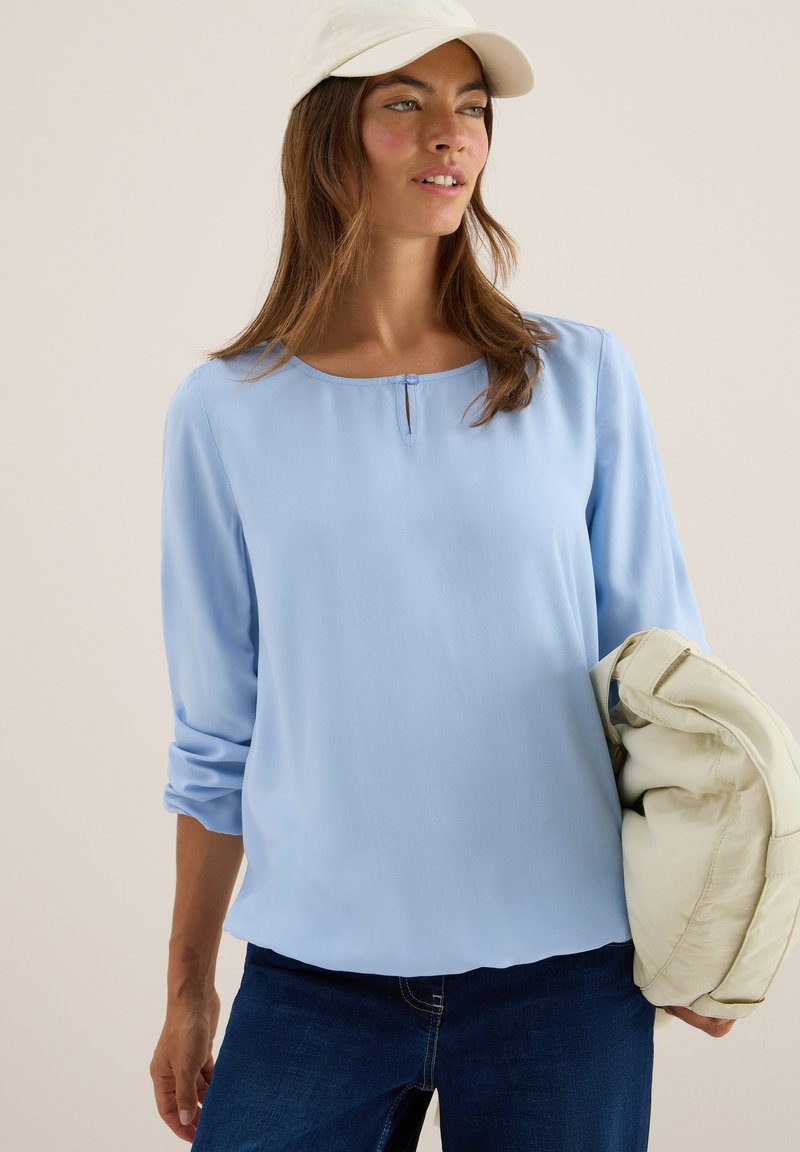Lichtblauwe blouse van glad materiaal met een ronde halslijn en een klein knoopdetail. Lange, opgerolde mouwen gecombineerd met donkerblauwe jeans.