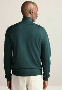 Tumši zaļa augstās apkakles džemperis ar mīkstu tekstūru, pieguļošu dizainu un garām piedurknēm, savienots ar gaišas krāsas biksēm.