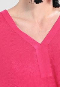 Primo piano di una persona che indossa un top V-neck testurizzato di un brillante colore fucsia e orecchini neri con frange.