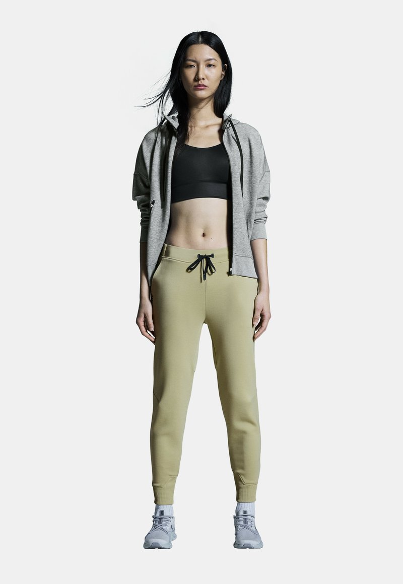 Gray zip-up hoodie, black sports bra, beige sweatpants with drawstring, and gray athletic shoes. Smooth fabric with fitted design.
